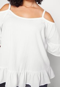 Top bianco scollato sulle spalle realizzato in tessuto testurizzato, con maniche corte e orlo a volant per un tocco di dettaglio.
