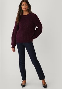 Vrouw staat gekleed in een gestructureerde donkerpaarse trui, donkerblauwe spijkerbroek en zwarte puntige hakken, poseert met één hand in de zak tegen een grijze achtergrond.