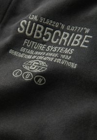 Svart tyg med broderad text i ljusgrått, som inkluderar "SUB5CRIBE" och geografiska koordinater, med en slät struktur och minimalistisk design.