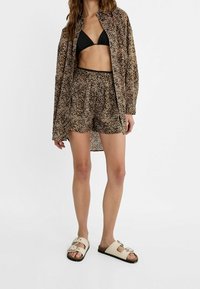 Kraťasy s leopardím potiskem a odpovídající volná košile, oboje v odstínech hnědé a černé. Černý bikinový top spárovaný se světle zbarvenými sandály.