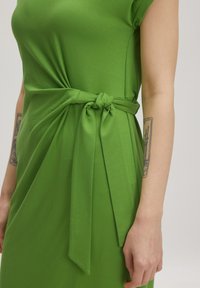 Grünes Kleid aus glattem Stoff mit einem geknoteten Band in der Taille. Kurze Ärmel und ein drapiertes Design sorgen für eine figurbetonte Silhouette.