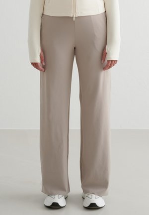 Persoon draagt beige wijde broek, witte zip-up top met duimgaten, en witte sneakers, staand tegen een lichte achtergrond.