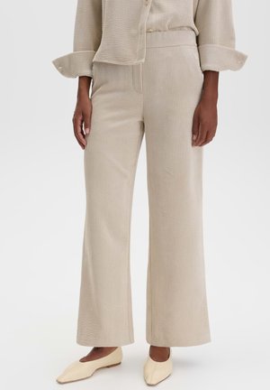 Persona che indossa pantaloni beige a gamba larga a coste con top a manica lunga abbinato e scarpe basse color crema, in piedi davanti a uno sfondo semplice.