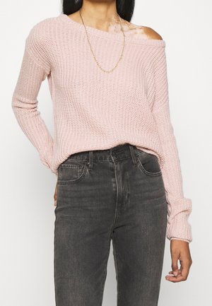 Vrouw draagt lichtroze off-shoulder gebreide trui, zwarte high-waisted jeans en een dunne gouden ketting, staand met één hand in de zak.