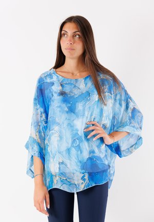 Blusa trasparente e fluida con un motivo a marmo astratto blu. Collo rotondo, maniche larghe e orlo scallopato. Abbinata a pantaloni scuri.