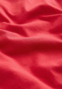 Červená bavlnená tkanina s hladkou textúrou, zobrazujúca jemné záhyby a vrásky, ktoré zdôrazňujú jej mäkkosť a pružnosť.