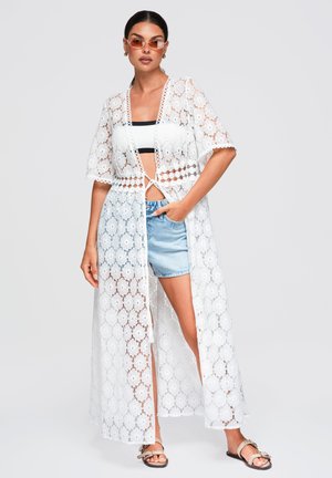 Donna che indossa un lungo kimono bianco in pizzo, un top corto bianco e nero, pantaloncini di denim, sandali beige e occhiali da sole arancioni, in piedi davanti a uno sfondo semplice.