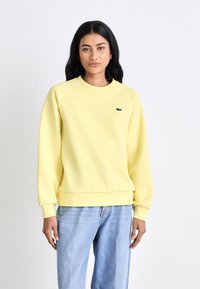 Sweatshirt amarelo feito de tecido macio, com decote redondo, punhos e barra canelados. Pequeno logótipo escuro no lado esquerdo do peito. Corte casual.