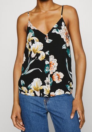 Kvinde iført en sort ærmeløs knappet top med stort gult og orange blomsterprint og blå jeans.