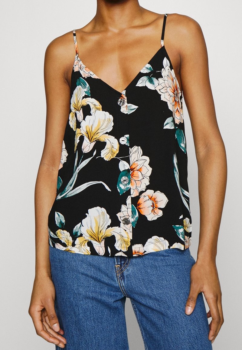 Femme portant un haut noir sans manches boutonné avec un grand imprimé floral jaune et orange et un jean bleu.