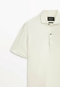Polo shirt blanc à manches courtes, fabriqué en tissu coton lisse, avec une patte de boutonnage à deux boutons et un col classique avec une texture subtile.
