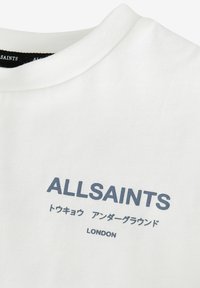Bílá bavlněná tričko s kulatým výstřihem, horizontální žebrovanou strukturou a šedým textem "ALLSAINTS" a "LONDON" napsaným různými fonty.