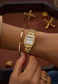 Zlaté náramkové hodinky so štvorcovým ciferníkom a rímskymi číslicami, osadené na leštenom, prepojenom remienku. Na zápästí je hladký zlatý náramok.