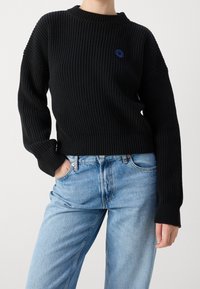 Pull en maille côtelée noire avec un col rond et un logo smiley bleu, associé à un jean taille haute bleu clair.
