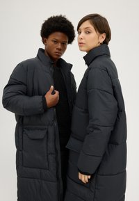 Two young adults wearing long black puffer jackets, standing close together against a plain light background.