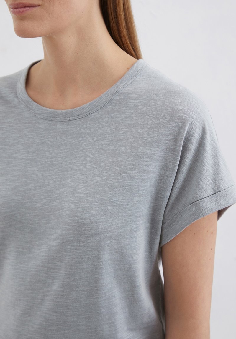 Hellgrau T-Shirt aus weichem, strukturiertem Stoff, mit Rundhalsausschnitt und aufgerollten kurzen Ärmeln. Legere, lockere Passform.