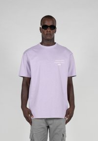 Lavender cotton t-shirt with short sleeves, crew neck, and white printed text on the front. Styled with gray cargo shorts and black sunglasses.