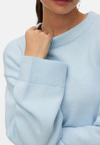 Pull tricot bleu clair avec encolure et poignets côtelés. Gros plan sur la texture du tissu et la manche pliée. Main de couleur peau serrée doucement.