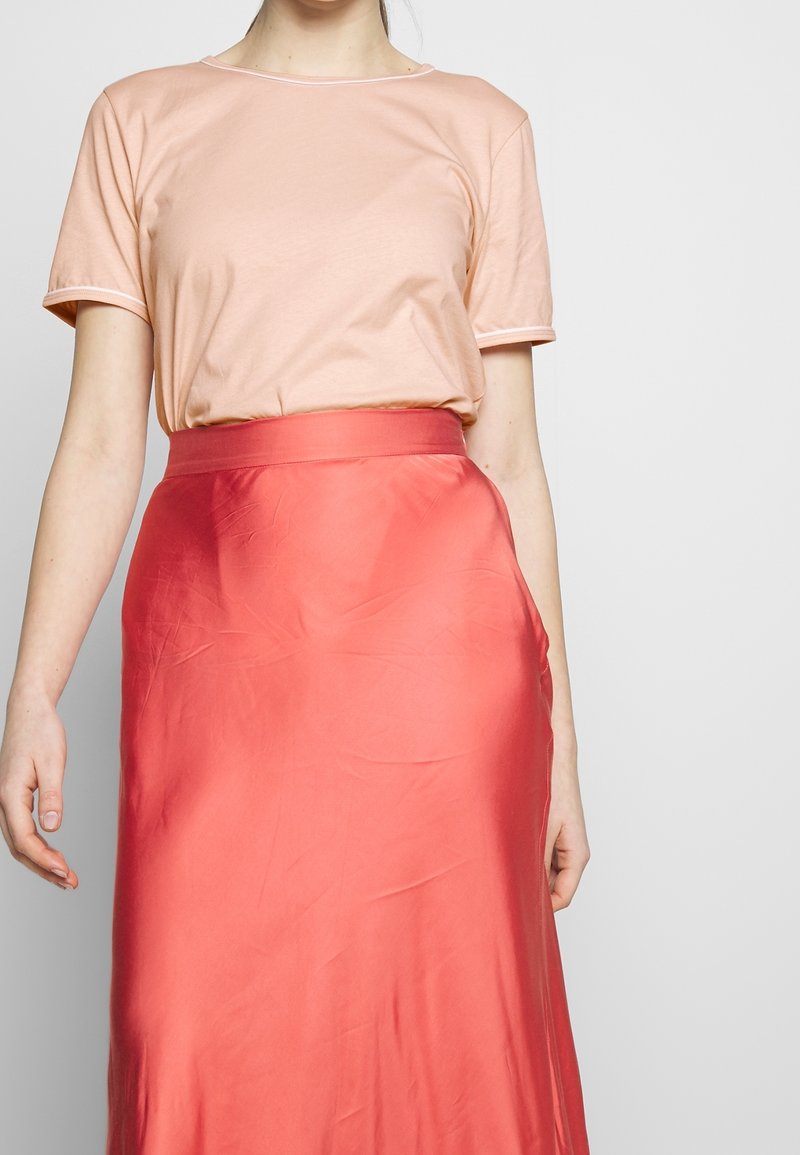 Personne portant un t-shirt rose pâle à manches courtes rentré dans une jupe midi corail brillante taille haute, debout devant un fond uni.