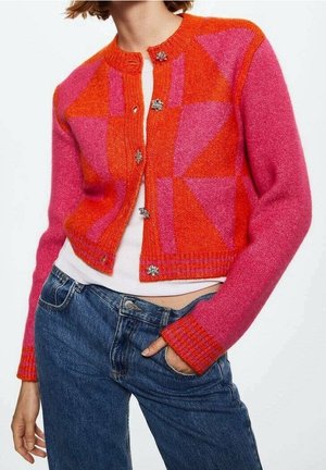 Femme portant un cardigan rose et orange à motif géométrique avec des boutons fleuris, une chemise blanche et un jean bleu.