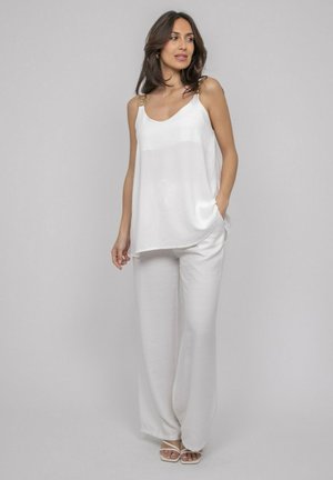 Femme debout portant un haut blanc ample sans manches avec des bretelles décoratives, un pantalon large assorti et des talons blancs ouverts, devant un fond uni.