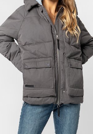 Woman wearing gray padded winter jacket with large front pockets and zipper, paired with blue jeans, hands in jacket pockets.