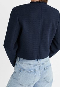 Veste texturée bleu marine avec un design court et un col effiloché, associée à un jean taille haute bleu clair, avec des poches et des détails de couture visibles.