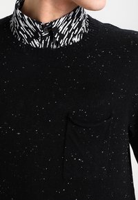 Uomo che indossa un maglione a macchie nere con una tasca frontale sopra una camicia a collo con pattern in bianco e nero.