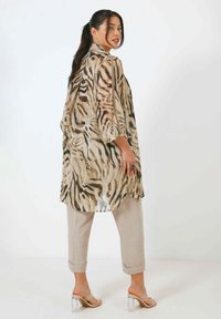 Langes, leichtes, durchscheinendes beiges Hemd mit braunem Zebra-Muster, kombiniert mit hellbeigen, verkürzten Hosen und klaren Sandalen mit Blockabsatz.