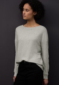 Světlý šedý svetr s dlouhým rukávem, kulatým výstřihem a lehce oversized střihem. Bavlněná směs s žebrovanými manžetami. Nošený s černými džínami.