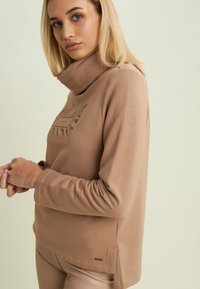 Pullover a maniche lunghe color beige con colletto alto, caratterizzato da un design testurizzato sulla parte frontale e fessure laterali per la ventilazione, realizzato in un materiale morbido.