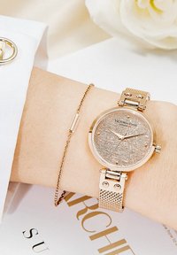 Orologio in oro rosa con quadrante testurizzato, cinturino a maglia e indicatori delle ore delicati. Accompagnato da un bracciale sottile in oro su un polso.