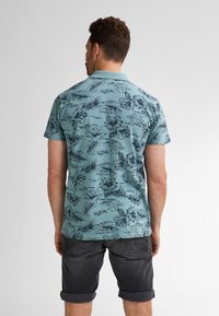 Pánský polo triko s krátkým rukávem v barvě teal se tmavými květinovými a listovými vzory. Má límeček a uvolněný střih, doplněný o kraťasy s ohrnutým lemem.