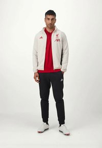 Light gray zip-up jacket with red Liverpool logo, red shirt, and black joggers featuring white Adidas stripes and white athletic shoes.