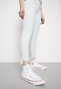 Bílé vysoké tenisky s gumovou podešví, kovovými očky a červenými a modrými akcentovými pruhy. Nosené s světlými modrými úzkými džíny se roztrhanými lemy.