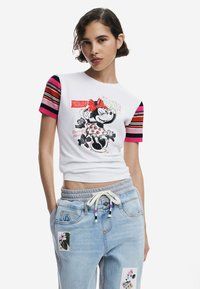 Witte cropped t-shirt met grafische print van een karakter. Beschikt over gestreepte roze en bordeauxrode mouwen. G gedragen met lichtblauwe broek versierd met patches.