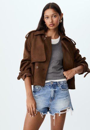 Giacca cropped in suede marrone con maniche arrotolate, abbinata a una maglia grigia e pantaloni shorts in denim strappati. Presenta chiusura con bottone e una texture liscia.