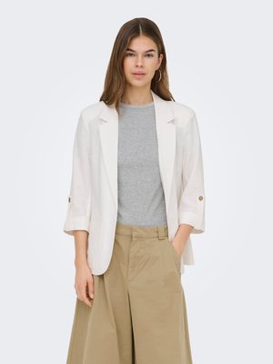 Jeune femme portant un blazer blanc sur une chemise grise et un pantalon beige à jambes larges, debout avec une main dans la poche devant un fond uni.