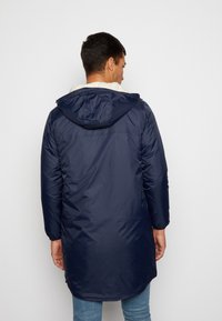 Marineblå puffjakke med hette, som har en myk hvit fleecefôring og lange ermer. Designet har en litt lengre bakhem.