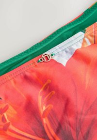 Blommönstrad tyg med dominerande nyanser av orange och grönt, med en grön dragkedja och en silverfärgad cirkulär detalj nära stängningen.