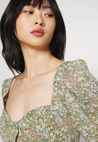 Blusa floral em verde com mangas curtas e bufantes. Apresenta um decote quadrado e detalhe de botões, adornada com pequenas flores rosas e azuis.