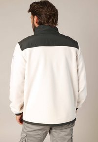 Homme avec des cheveux bruns et une barbe portant une veste en polaire blanche avec des panneaux d'épaule et un col gris foncé, et un pantalon cargo gris clair.