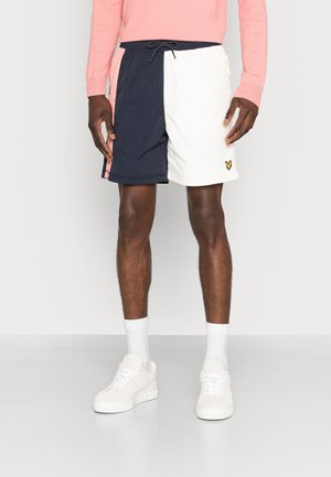 Mand iført shorts i navy og hvid blokfarve, lyserød langærmet top, hvide sokker og hvide sneakers, stående mod en hvid baggrund.