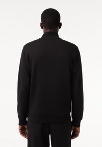 Uomo visto da dietro che indossa una giacca nera a maniche lunghe e pantaloni neri abbinati su uno sfondo bianco uniforme.