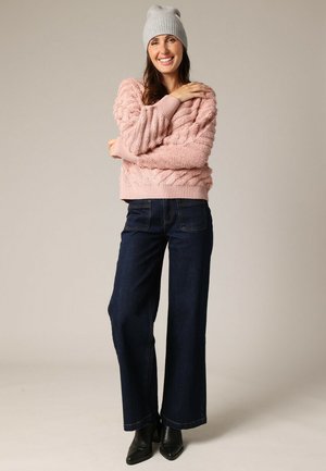 Pull en maille torsadée rose, jean foncé à jambes larges et bottines noires. Le modèle porte un bonnet gris clair et se tient les bras croisés.