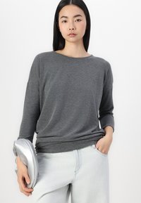 Grå långärmad topp i ett mjukt, texturerat tyg med en avslappnad passform. Matchad med ljusblå jeans och håller en silverfärgad clutch.