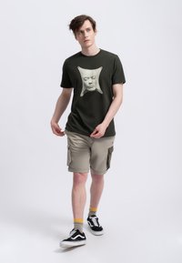 Donkergroene katoen t-shirt met een frontale grafiek van een gezicht, gecombineerd met beige cargo shorts en zwarte sneakers met witte zolen.