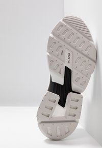 Suela de zapatilla blanca con características de goma texturizada, surcos prominentes y un acento negro en el centro para estabilidad y agarre.