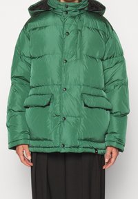 Veste rembourrée verte avec une finition brillante, dotée d'un col haut, de fermetures à boutons, de deux poches avant et d'un design ajusté et oversize.