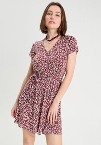 Robe bordeaux avec un petit imprimé floral blanc, manches courtes, décolleté en V, taille cintrée et jupe fluide. Accentuée d'une ceinture nouée.